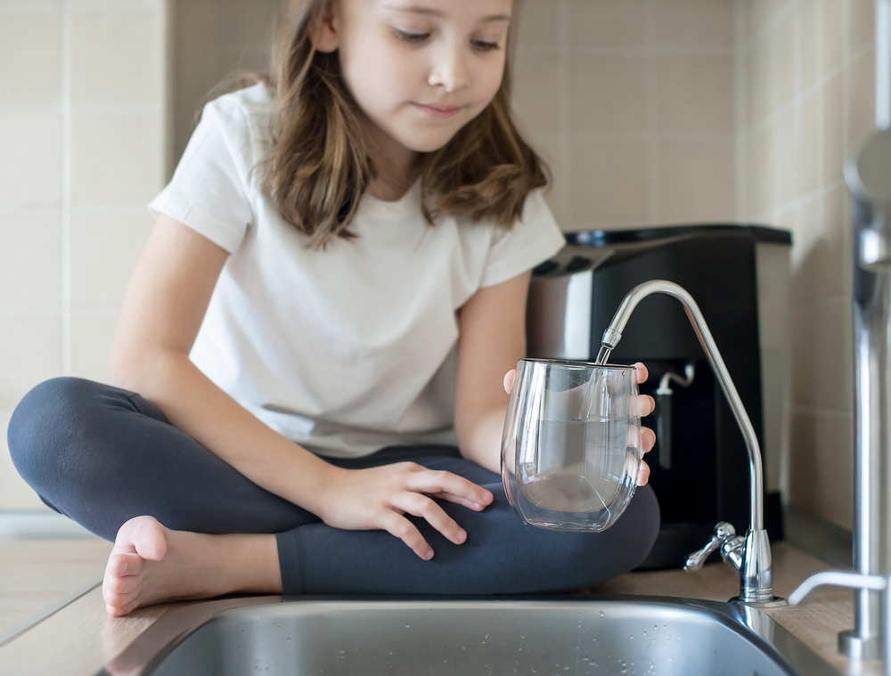 a young child sitting on the counter as she fills a glass of water from the kitchen sink