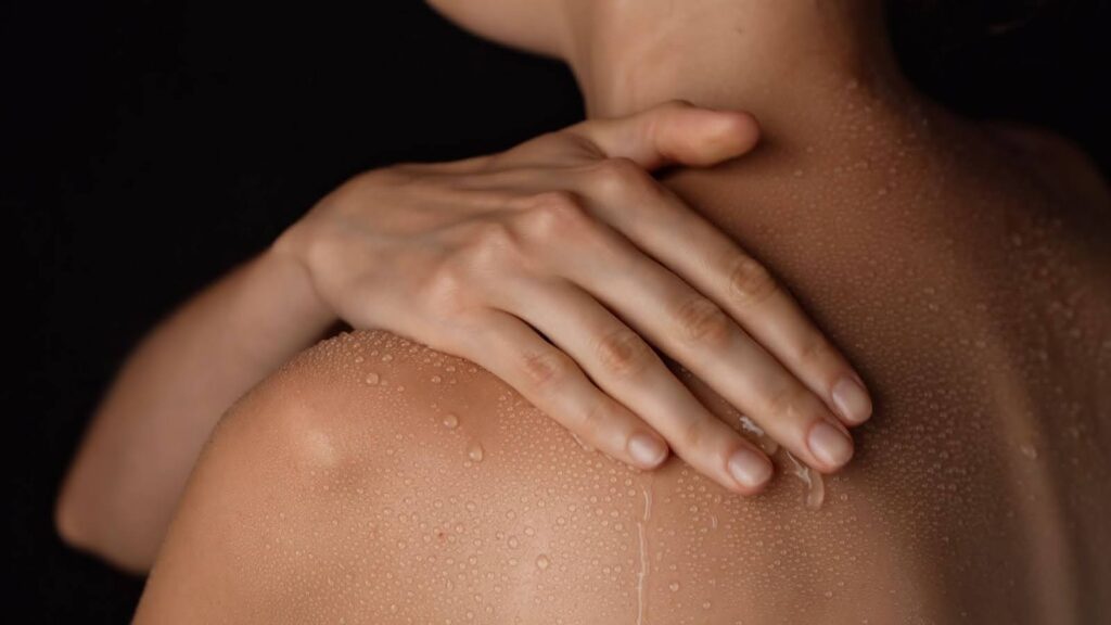 Close-up of a woman's shoulder with water droplets running down her back as she rubs her hand on her shoulder