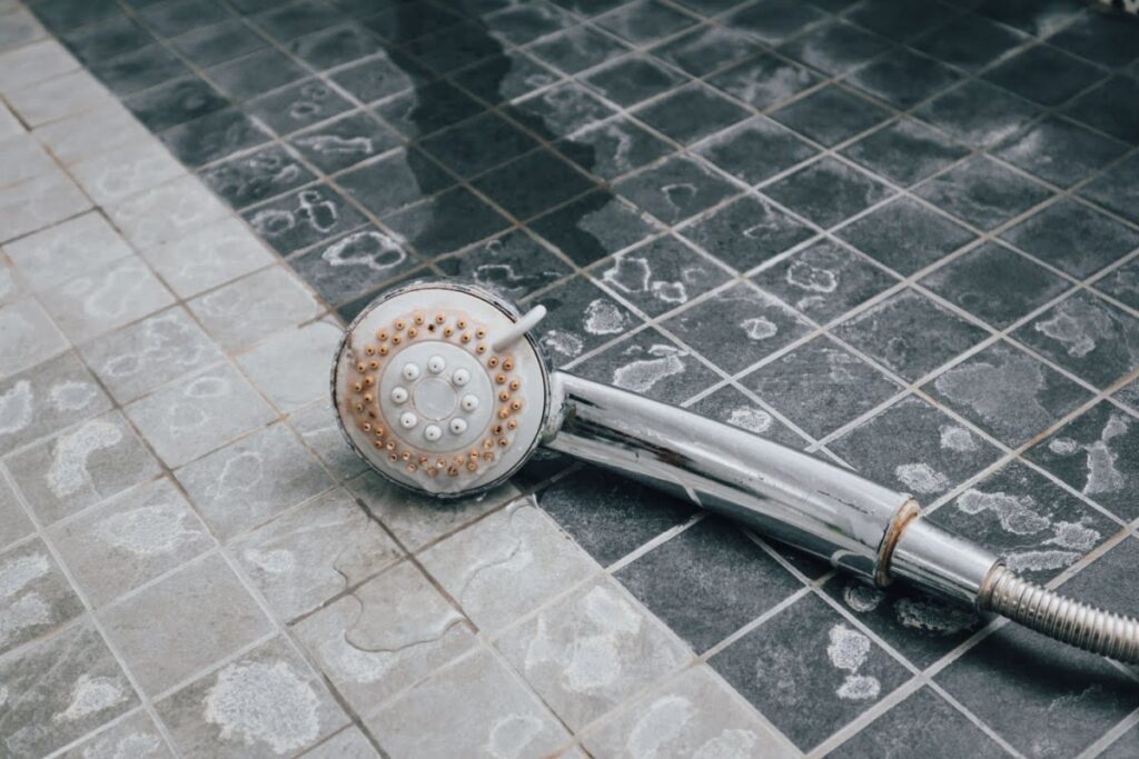 A detachable showerhead resting on the floor of a tiled shower with hard water stains covering everything