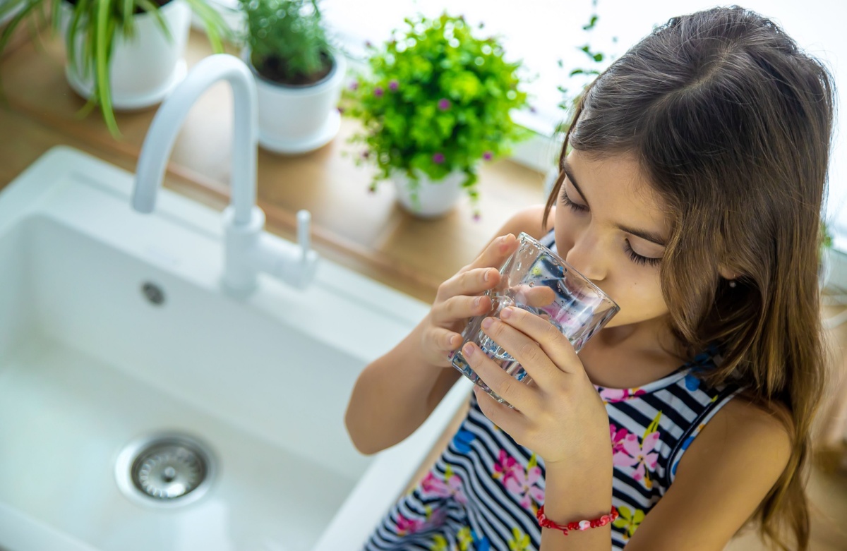 A young girl sitting on the counter next to the kitchen sink as she drinks a glass of tap water
