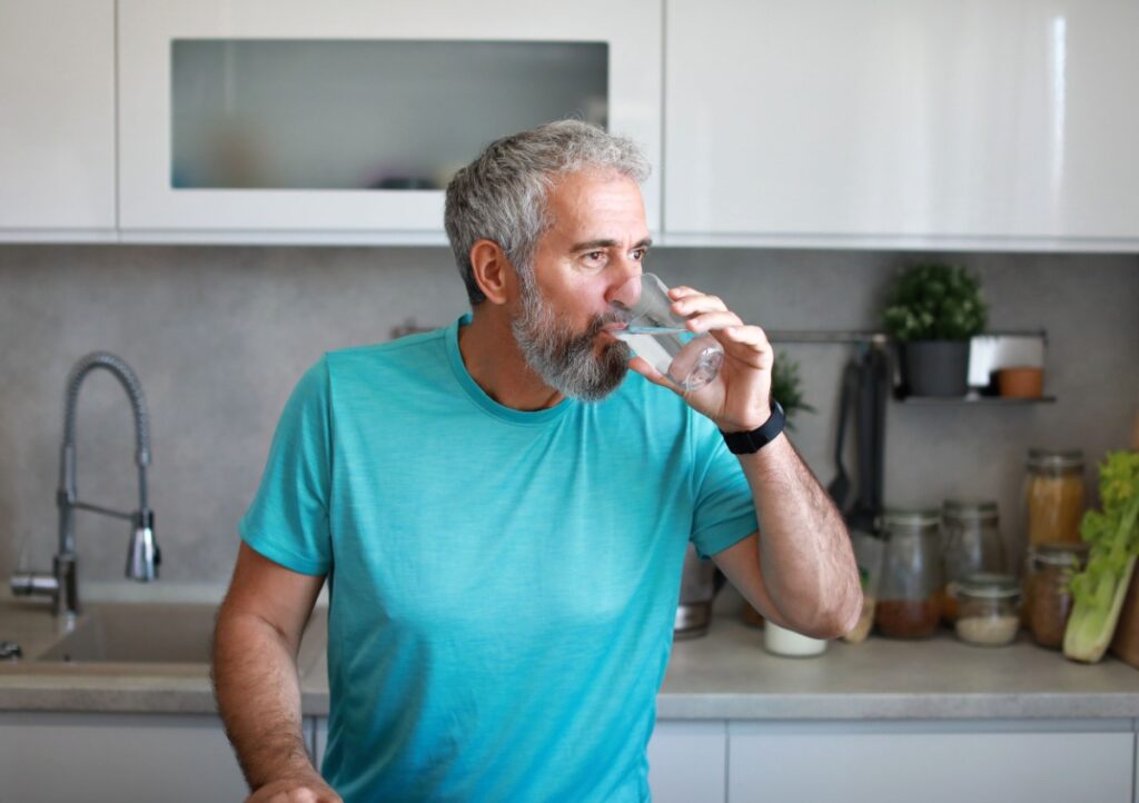 Man with gray hair and beard is drinking a glass of purified water in front of his kitchen sink after a workout